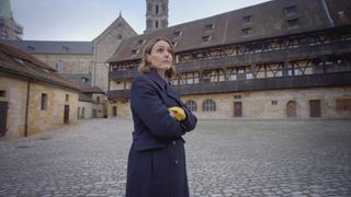 Suranne Jones stands in a courtyard in the Germany town of Bamberg. The buildings behind her are made of stone, with timber panelling.