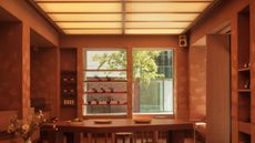 A softly glowing cafe interior clad in terracotta plaster with a textured surface, decorated with wooden furniture, high-tech sound systems, minimalist windows, and a back-lit, checkered ceiling.