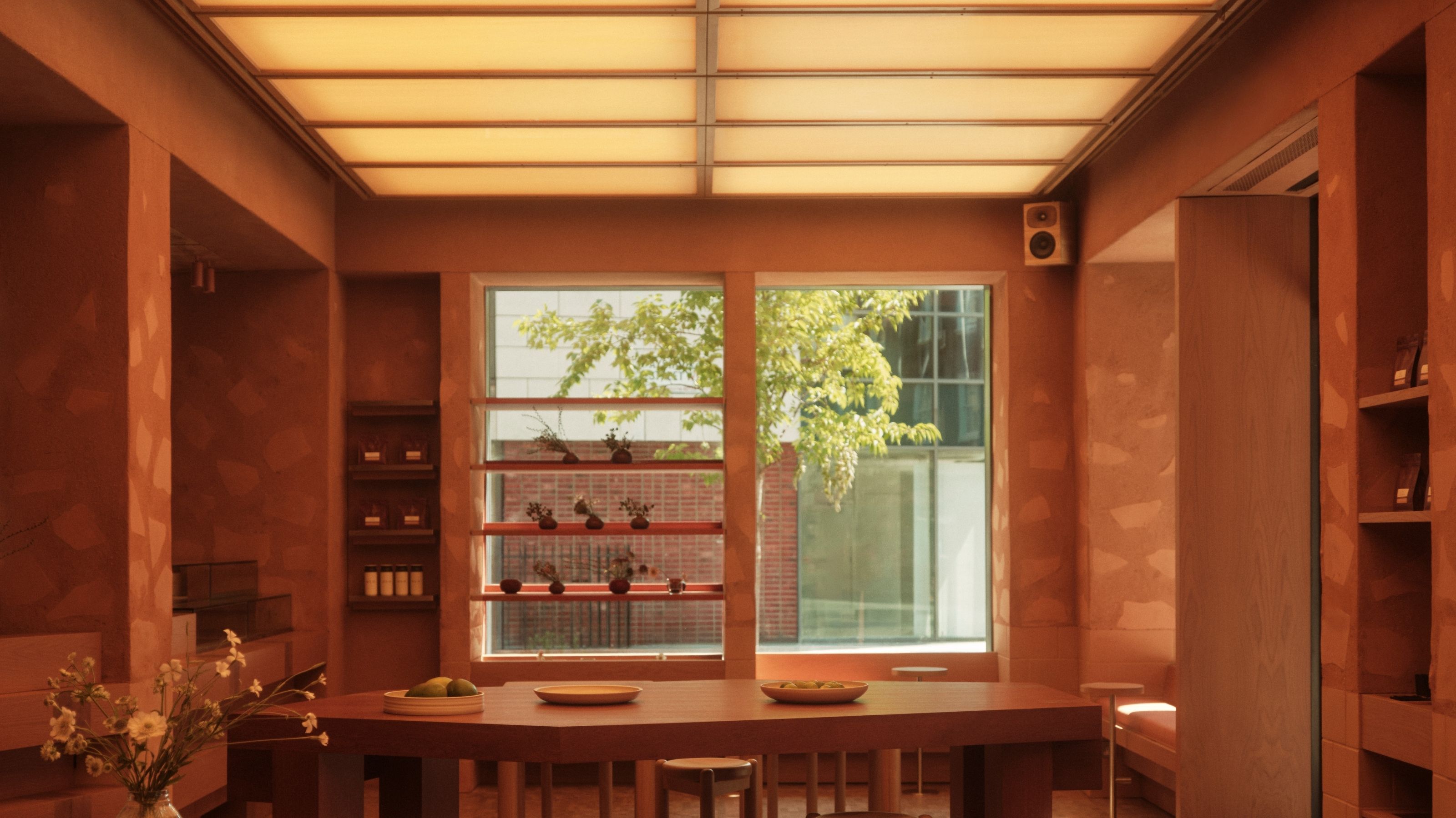 A softly glowing cafe interior clad in terracotta plaster with a textured surface, decorated with wooden furniture, high-tech sound systems, minimalist windows, and a back-lit, checkered ceiling.