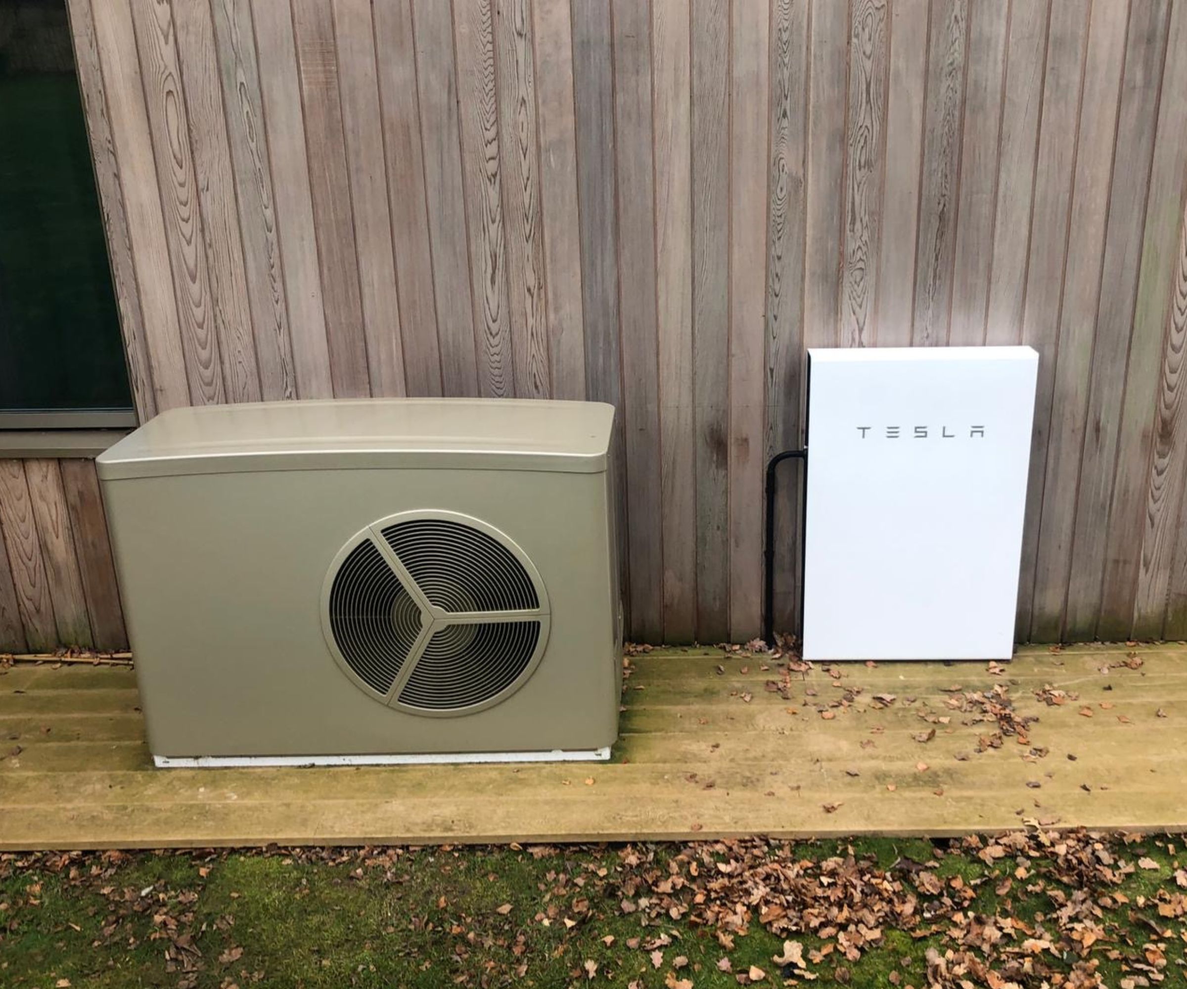Outdoor heat pump unit beside a Tesla Powerwall battery, installed on a timber deck against a wooden-clad house wall.