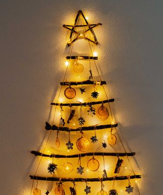 Handmade craft Christmas tree made from sticks and natural materials on a white wall with yellow lights illuminating it