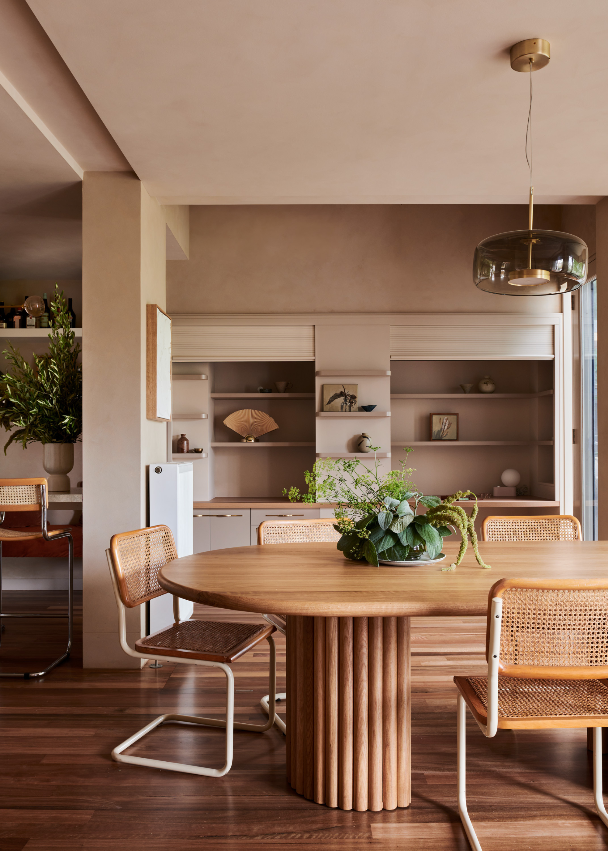A dining room with beige walls, a hanging glass pendant, a wood dining table with rattan style dining chairs, and a green centerpiece