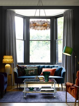 grey painted living room with bay window, blue sofa and armchair, orange armchair, floor lamp, table lamp, blue rug, modern chandelier, glass coffee table