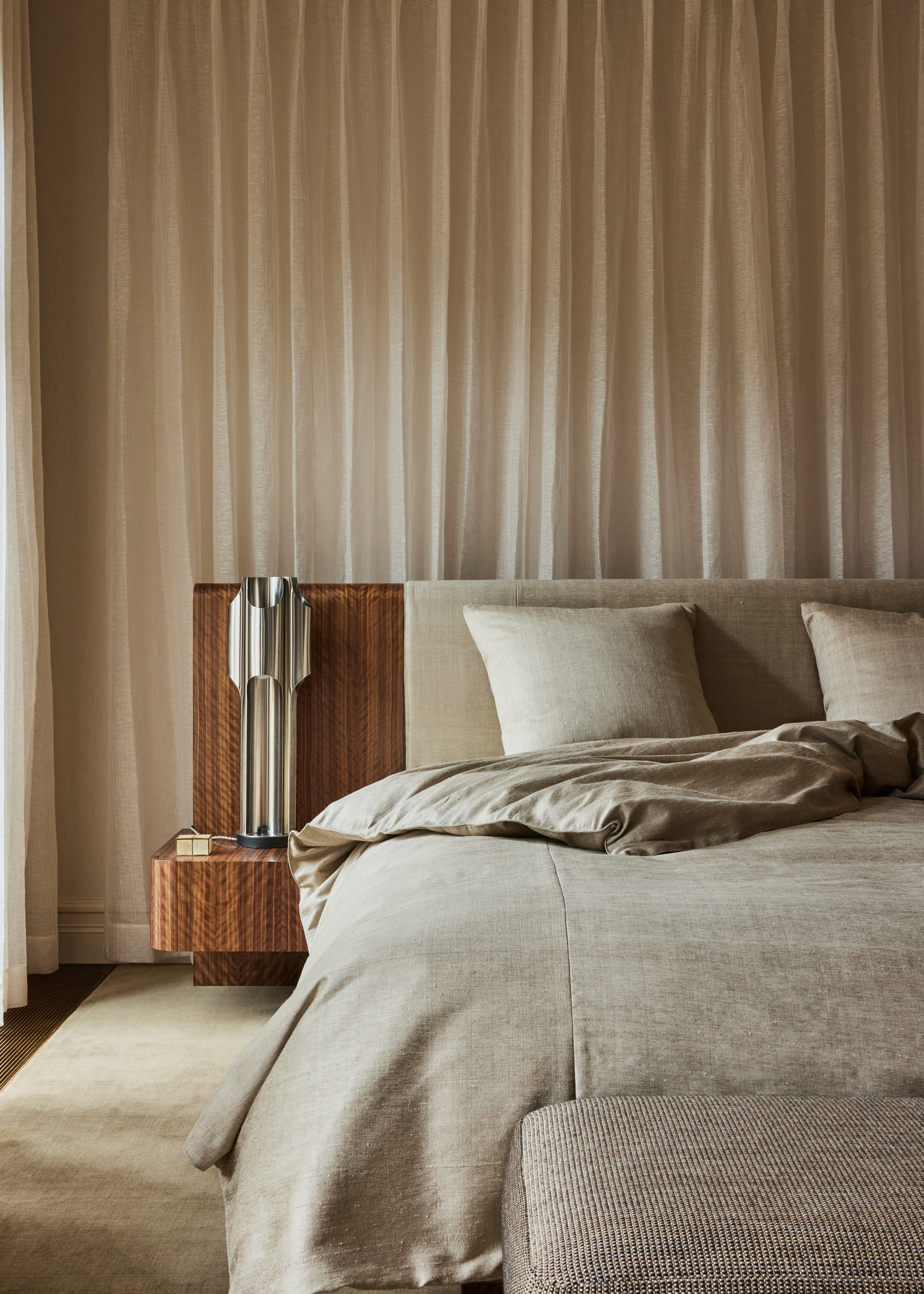 soft neutral bedroom with curtain behind bed, and built-in timber headboard with side tables and chrome table lamp