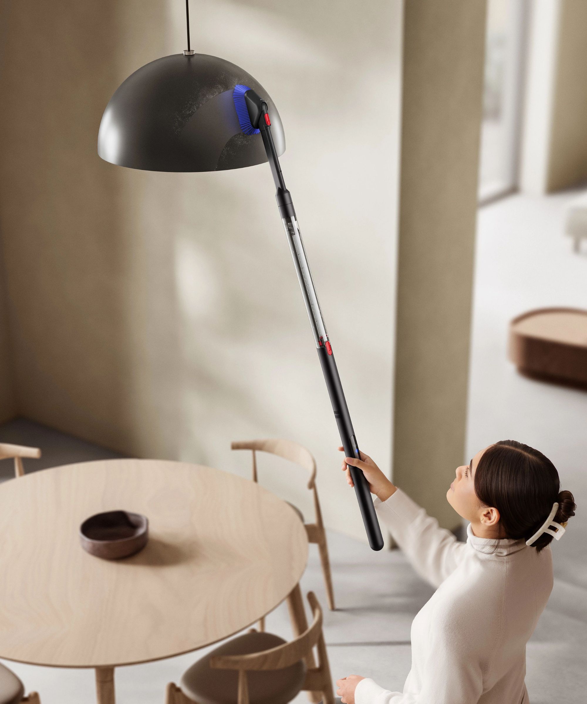 Dyson PencilVac cleaning a hanging light with dusting brush attachment, as person holds the vacuum above their head with one hand. Picture in the room are a wooden table and chairs, cream walls and white floors.