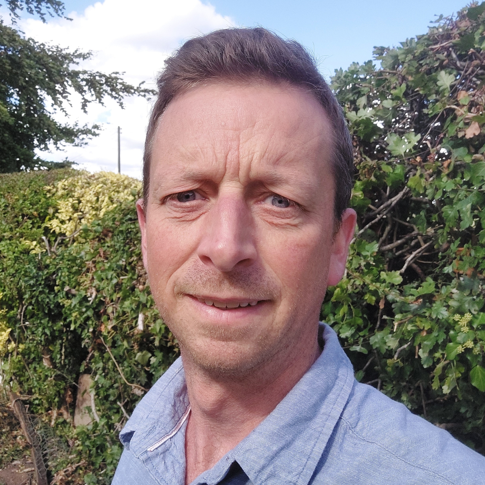 man with short hair wearing blue shirt stood with hedge in background