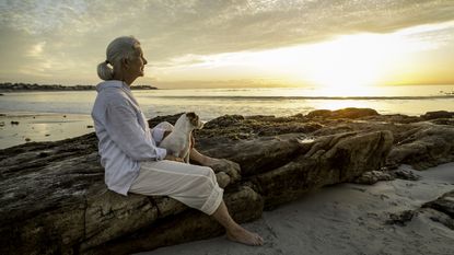 An older woman sits alone on a beach at sunset.