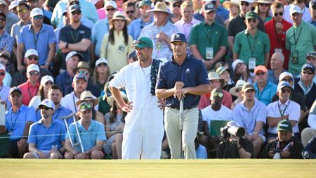 Bryson DeChambeau stands next to his caddie with putter in hand in front of a large number of patrons at The Masters in 2024