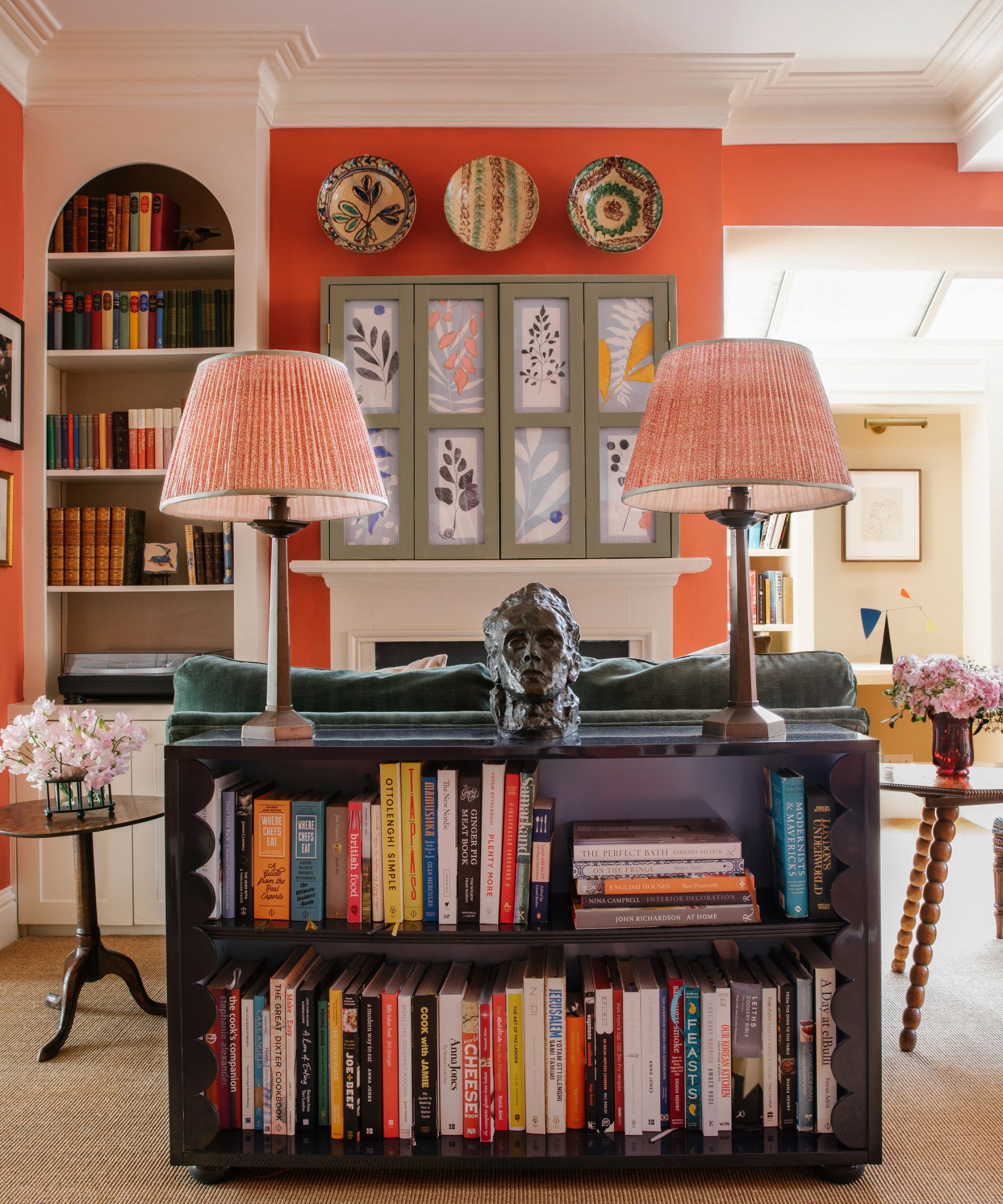A cozy living room styled in a grandma chic aesthetic, featuring coral walls, vintage bookshelves, a book-filled console table, patterned lampshades, and decorative plates displayed above a fireplace