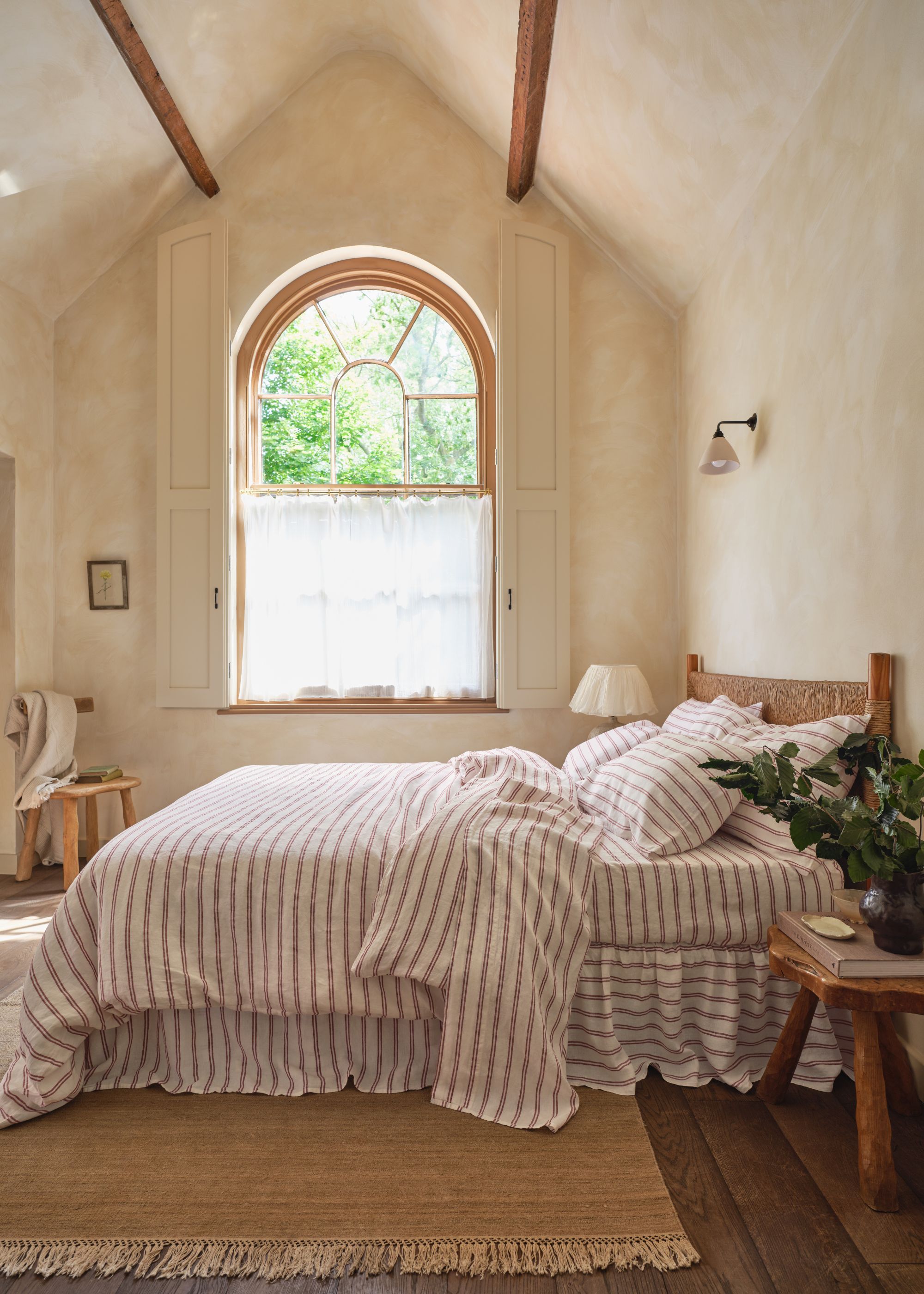 A modern rustic bedroom with sloped, high ceilings and an arched window on the far wall. Lime-wash warm white walls and a large king-sized bed with striped bedding.