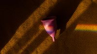 A pink silicon menstrual cup sits on a rubbery brown surface with a rainbow prism of light moving across it