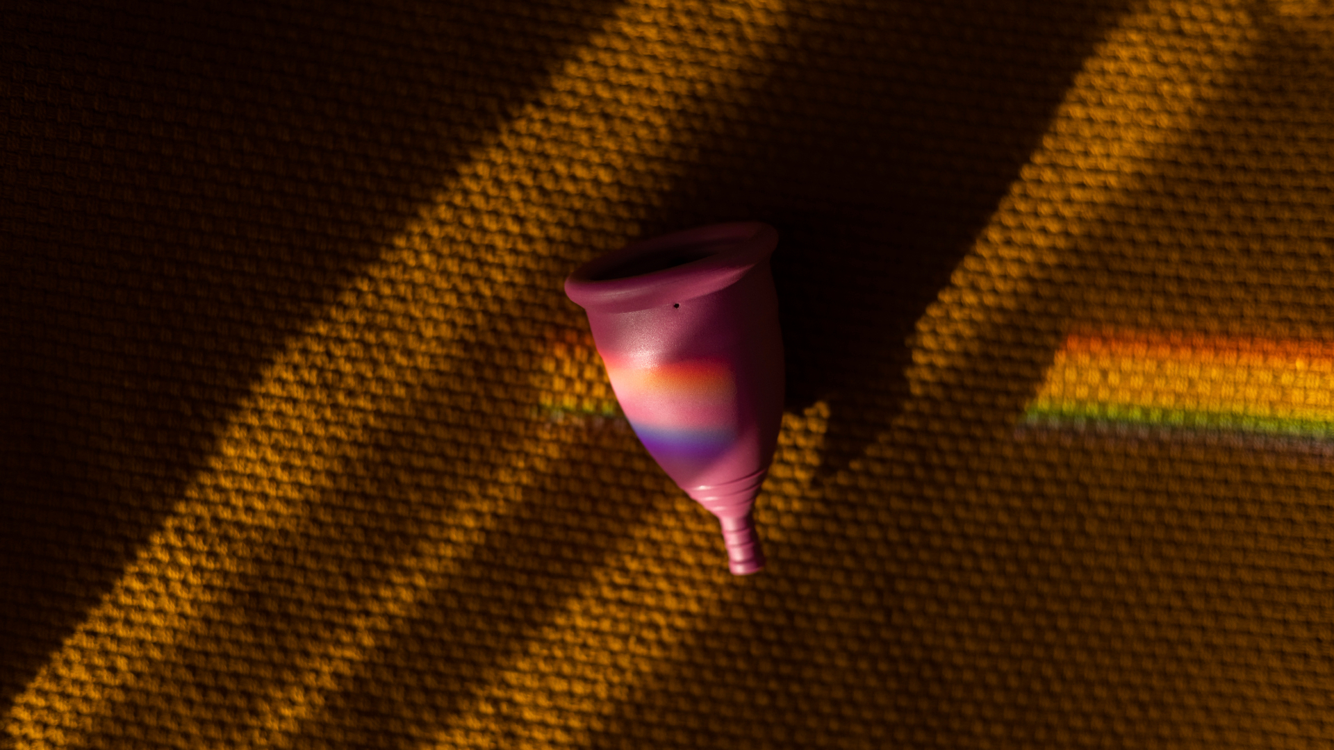 A pink silicon menstrual cup sits on a rubbery brown surface with a rainbow prism of light moving across it