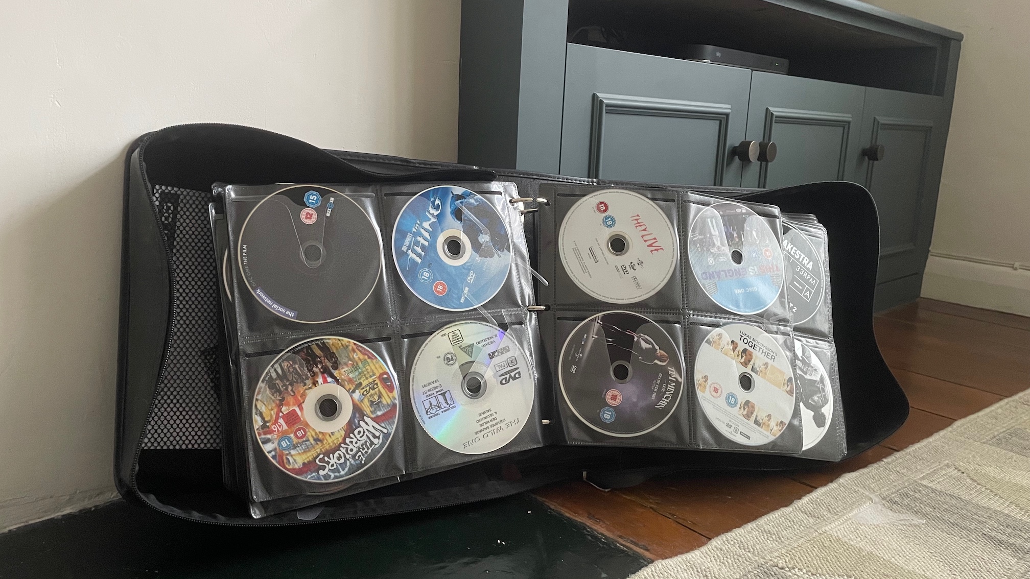 An open CD wallet full of DVDs, in front of a TV cabinet in a living area.