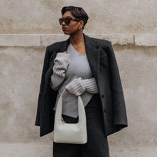 Influencer standing in front of wall wearing grey sweater, black blazer, and white Margaret Sherwood bag.