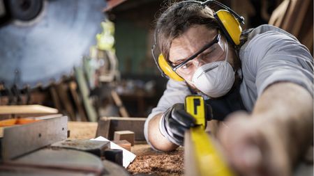 A man wearing a face mask and ear protection uses a tape measure on a length of board.