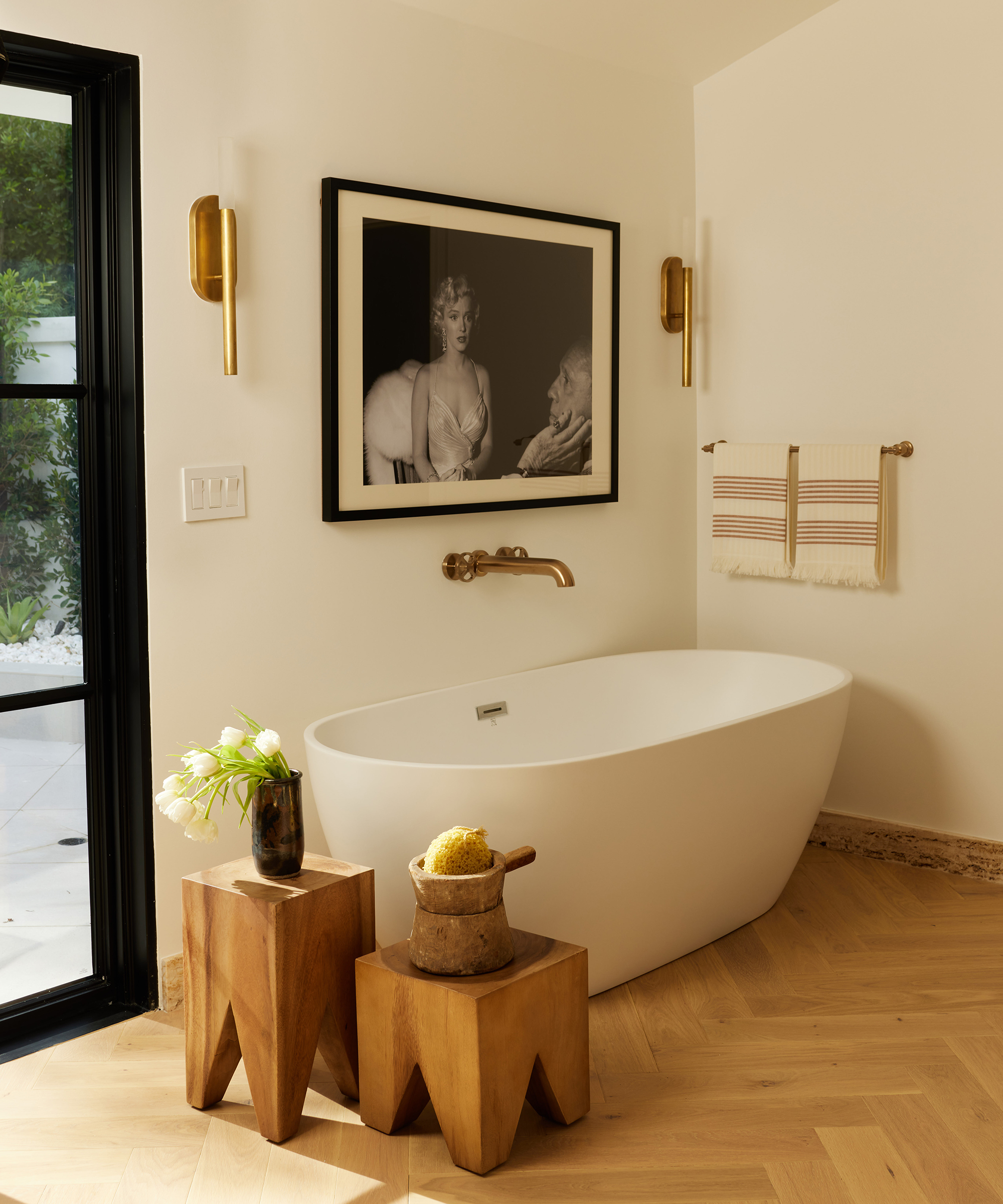 white bathroom with white bath and two wooden stools and a big framed piece of art on the wall