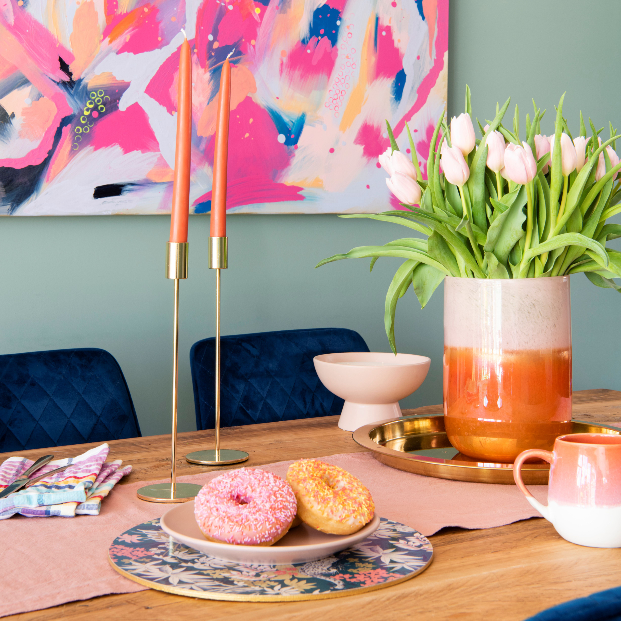 Wooden dining table with floral placemat, pink plate with doughnuts, gold candle holders and vase of tulips