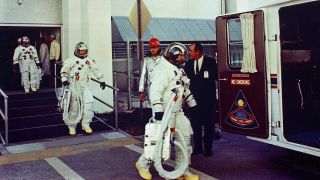 Three astronauts in their launch suits are walking out of a building and into a van.