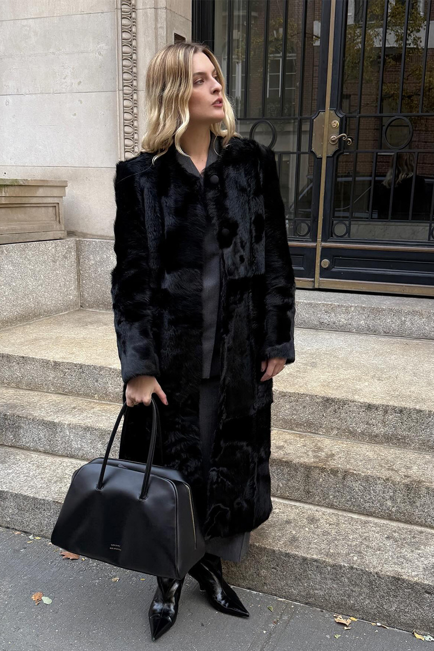 West Village winter trends are shown in a street-style photo of a woman standing in front of a glass doorway of a concrete apartment building in her winter outfit. She's wearing a long black fur coat over a black button-down shirt and skirt, styled with black pointed knee-high boots and a black bowler bag