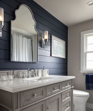 A bathroom with navy blue shiplap, an arched silver mirror, wall art and two silver sconces, a light gray sink unit with a white surface, candles, and a silver faucet and taps, and light gray patterned floor tiles