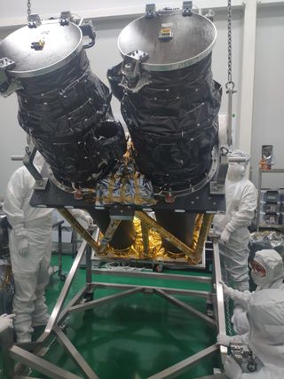 three humans dressed in full-body white coveralls work around a spacecraft in the shape of two large black cylinders held up by a metal frame.
