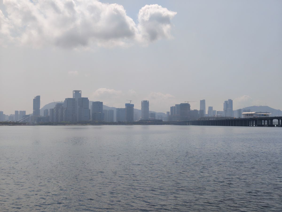 A wide landscape shot of a coastal city skyline under a hazy, overcast sky with several large, fluffy white clouds. A long bridge on the right extends across the water toward the distant skyscrapers. The water in the foreground is calm with small ripples, reflecting the muted light of the sky.