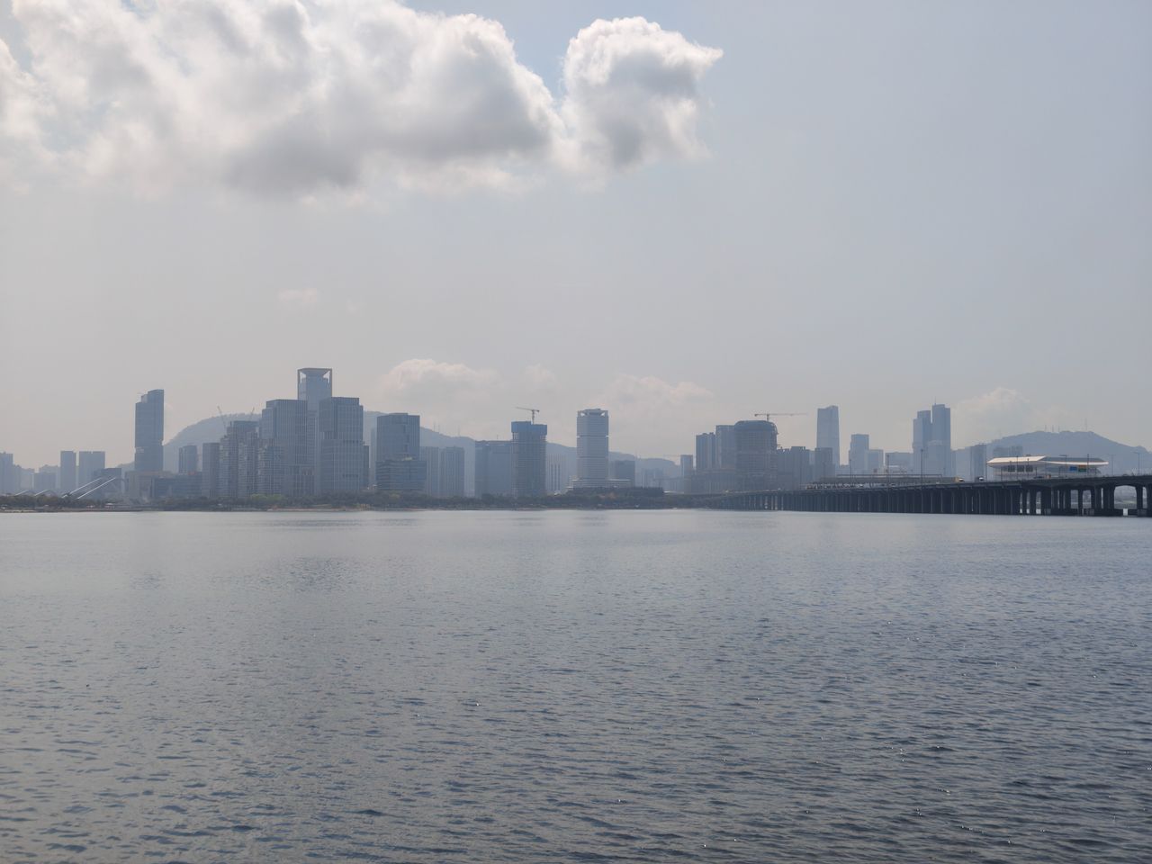 A wide landscape shot of a coastal city skyline under a hazy, overcast sky with several large, fluffy white clouds. A long bridge on the right extends across the water toward the distant skyscrapers. The water in the foreground is calm with small ripples, reflecting the muted light of the sky.