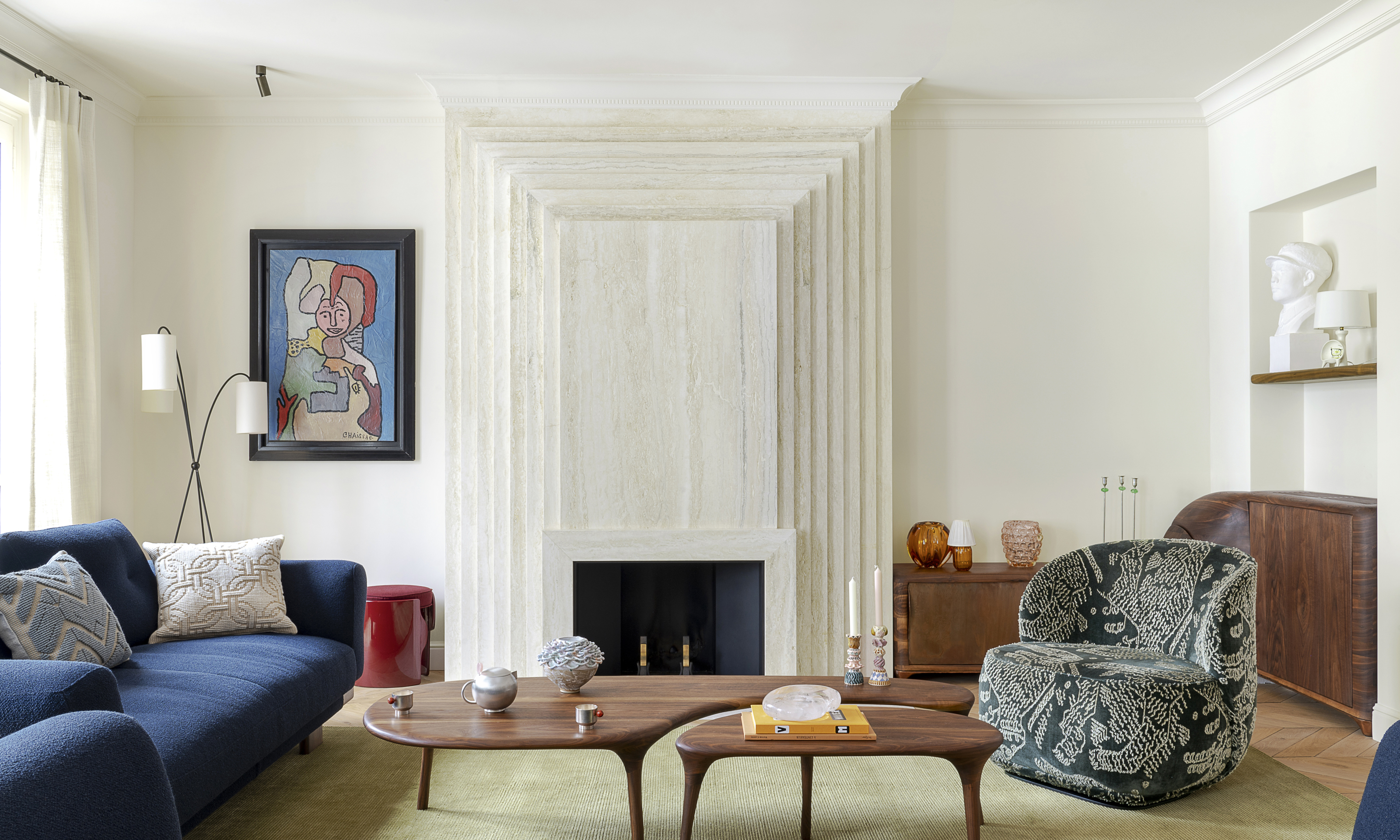 White living room with stone fireplace, pale green rug, dark blue sofa and sculptural wood coffee tables