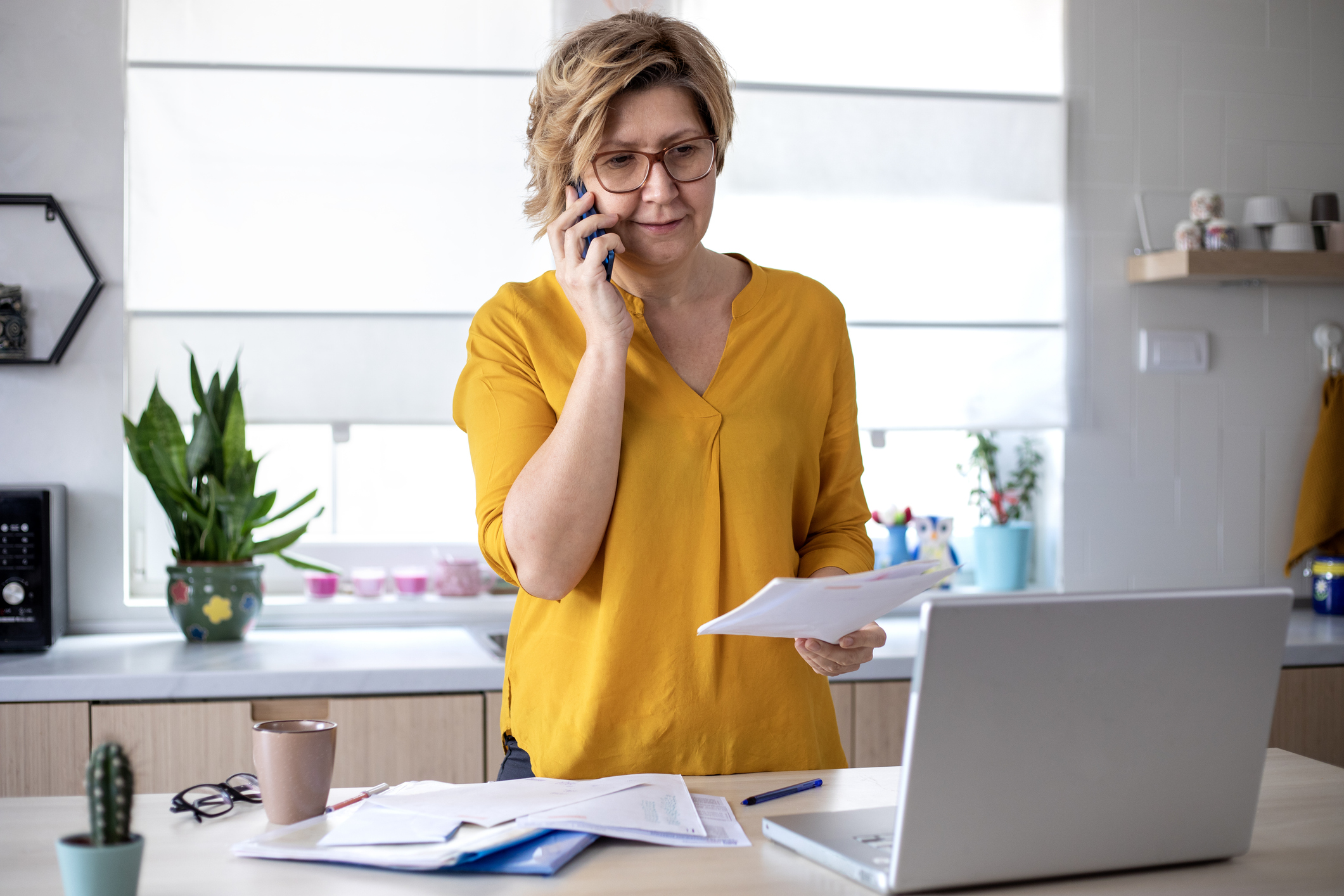 A woman stands in her kitchen, reviewing her finances while on the phone.