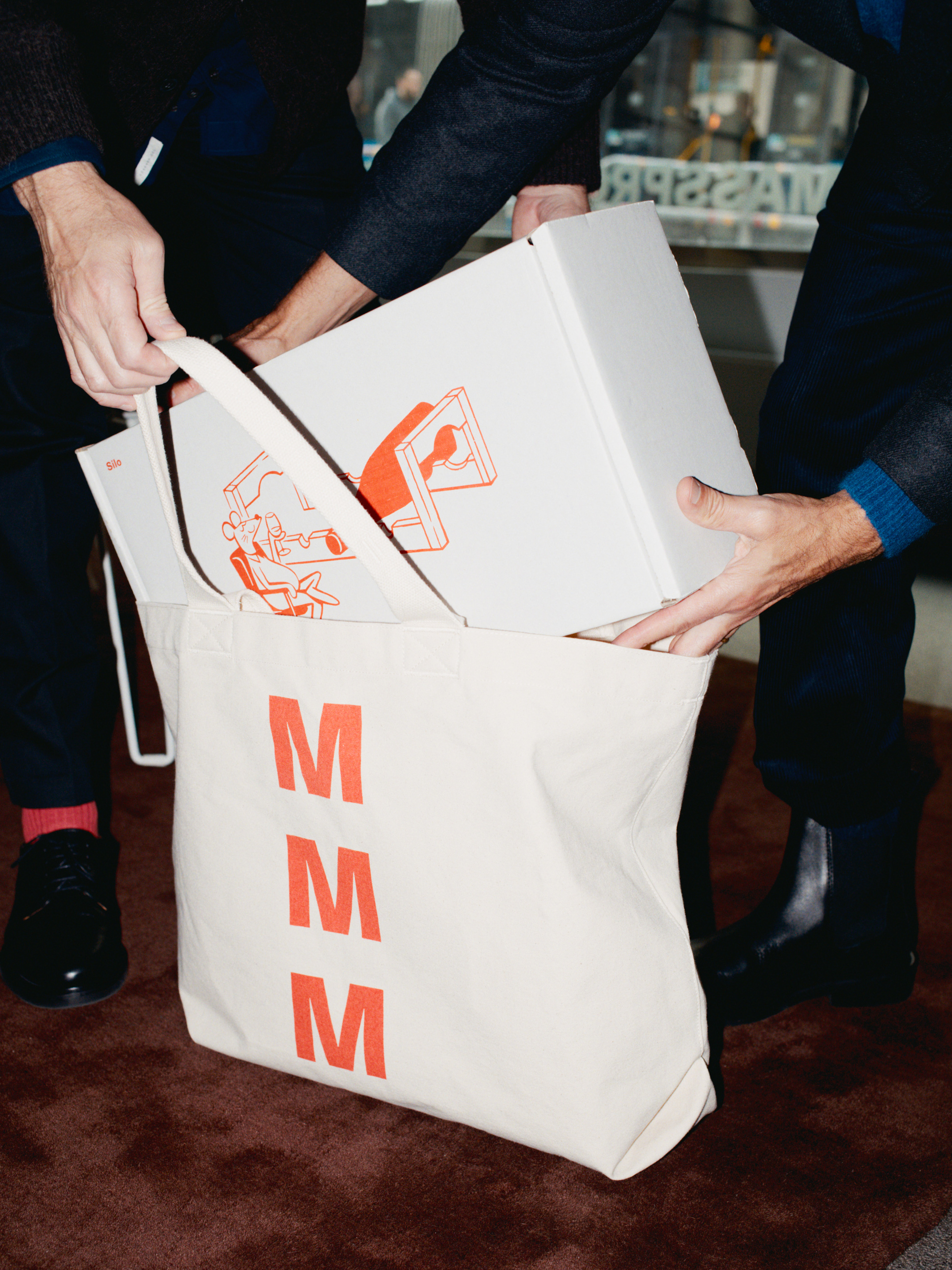 Image of two people in suits putting a white box with orange writing into a white bag with orange writing.
