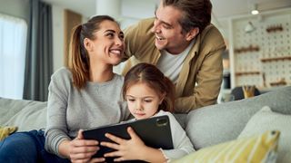 Happy parents spending time with their small daughter who is using a digital tablet while sitting on the couch in the living room.