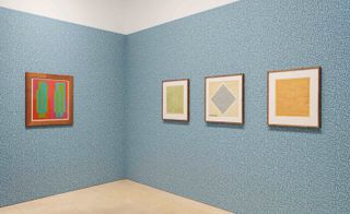 The corner of a room with 4 framed painting (one on the left and 3 on the right) featuring a white ceiling, light blue textured wall and beige tiled floor