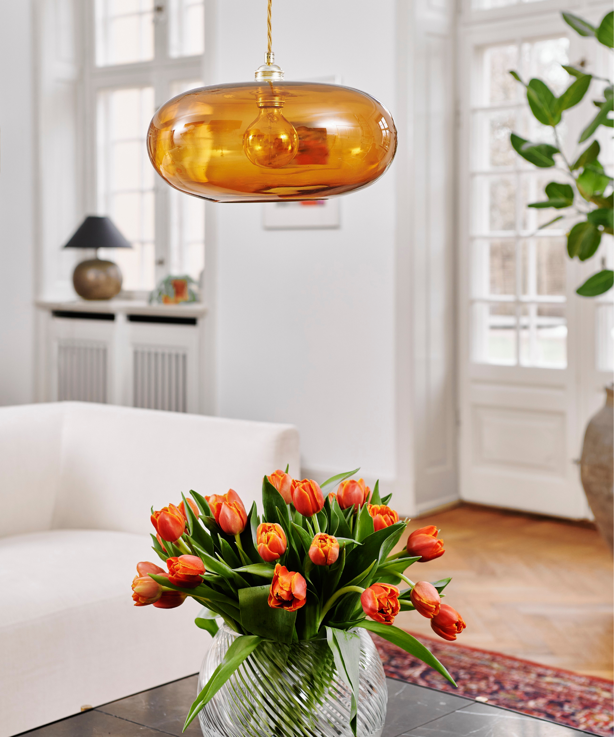 orange glass low pendant light hanging over vase or orange tulips on coffee table, cream sofa, table lamp on windowsill