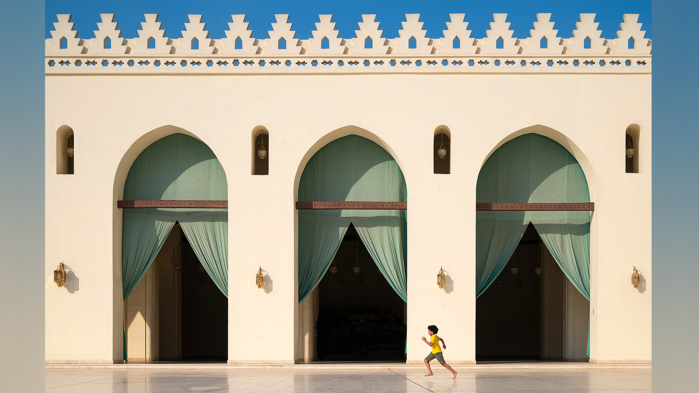 A young boy in a yellow shirt runs joyfully across a marble courtyard, framed by arched doorways with turquoise drapes