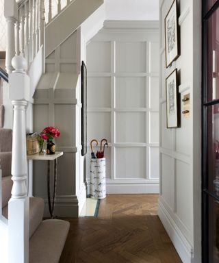 Off-white painted hallway with square paneling, warming wood parquet flooring, umbrella stand, carpeted staircase, framed artwork