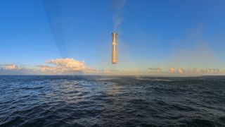 A giant metal cylinder hovers above a sea in front of a blue sky.