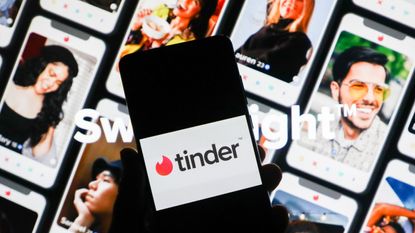 Tinder app logo is displayed on a mobile phone screen photographed on Tinder website background.