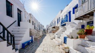 Image of a street in Greece lined with white square houses with blue accents. The sun is shining high in the sky.