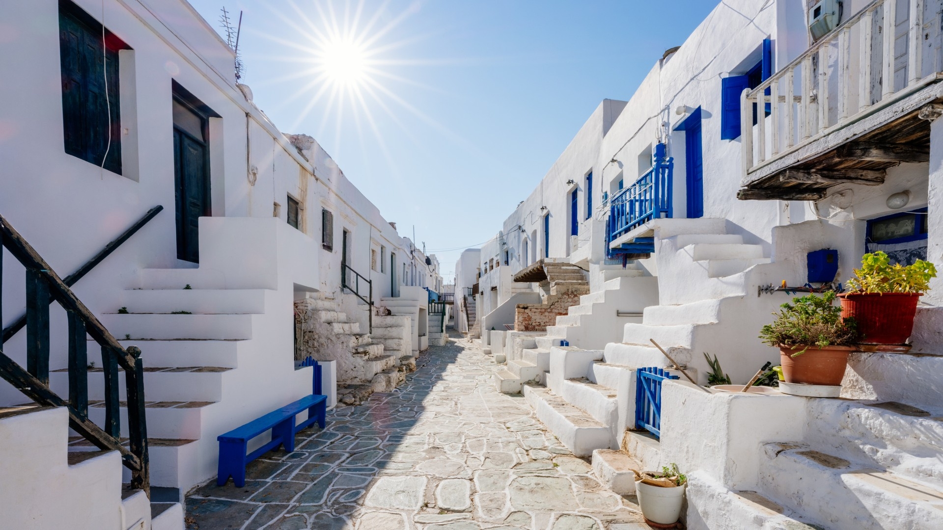 Image of a street in Greece lined with white square houses with blue accents. The sun is shining high in the sky.