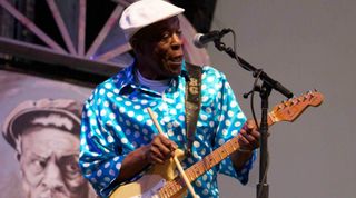 Buddy Guy photographed onstage playing a Fender Stratocaster electric guitar while playing the strings with a drumstick.