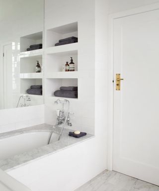 A white bathroom with shelves set into the wall, a door with gold hardware, and a bath with marble border