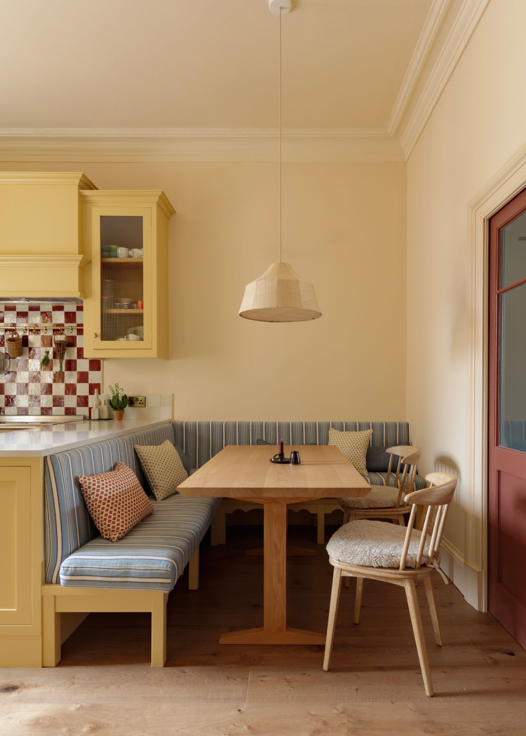 A modern kitchen with warm white walls, butter yellow cabinets, burgundy checkered backsplash tiles, and corner banquette seating with light blue striped upholstery.