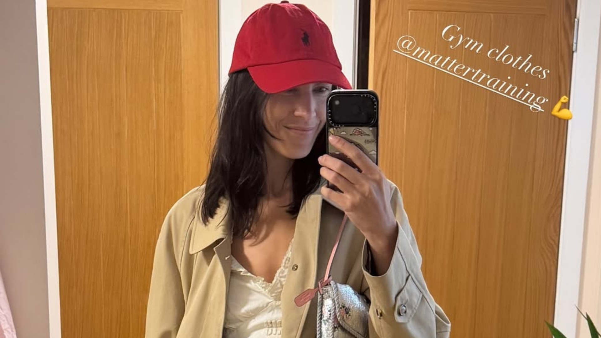 Alexa Chung takes a mirror selfie wearing a white cotton dress with a beige trench coat and a red cap.