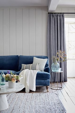 A living room with white shiplap and flooring, blue velvet sofa, faux marble coffee table and grey curtains