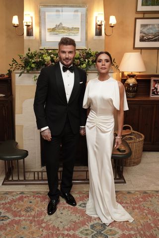 David Beckham wearing a black tux and Victoria Beckham wearing a white silik gown standing in front of a fireplace with flowers