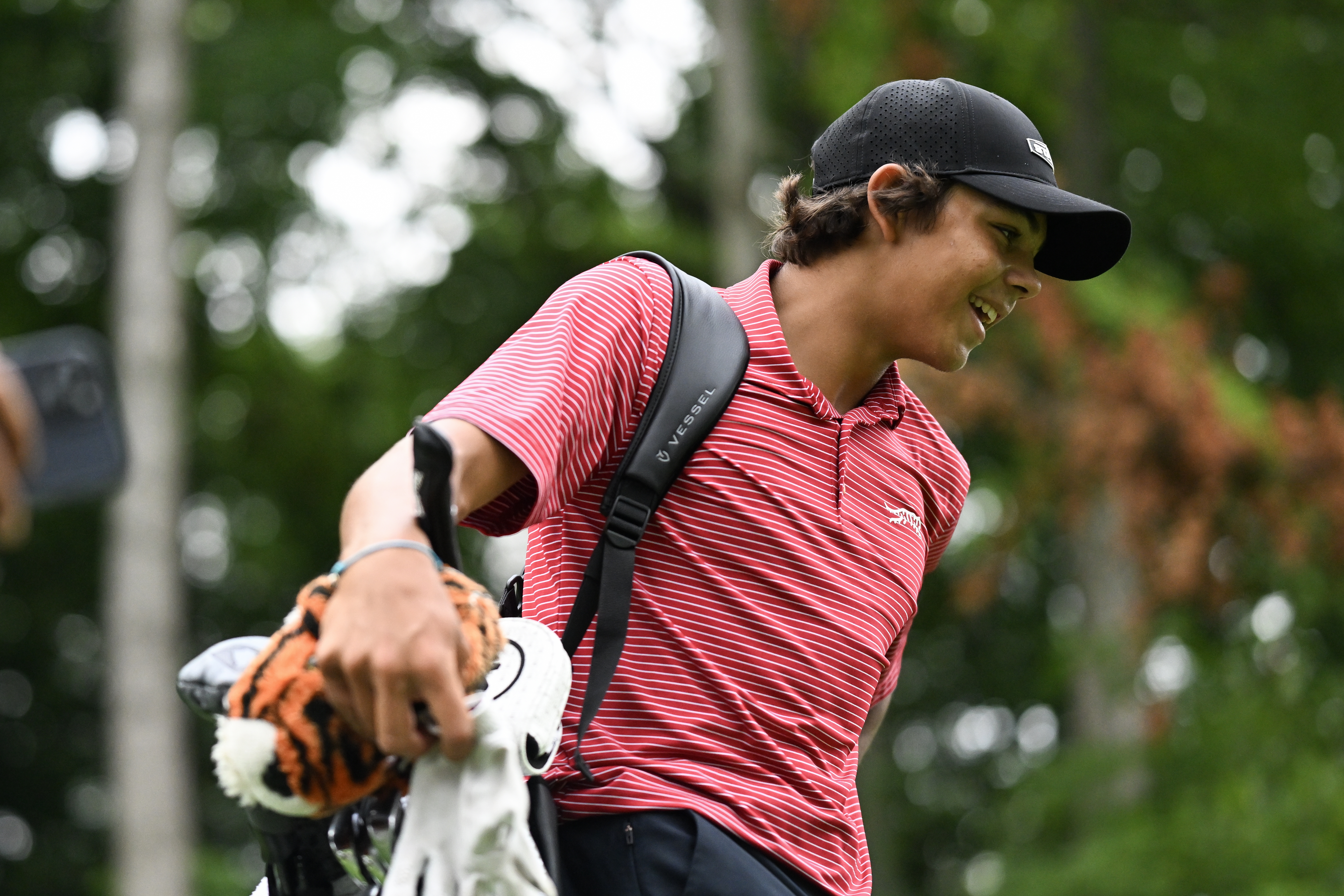 Charlie Woods smiles while carrying his golf bag in a red shirt and black cap