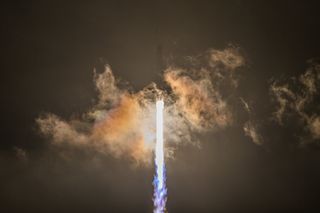 a white rocket lifts off at night above a plume of fire and smoke