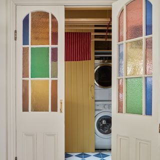 view into a utility room with multicoloured stained glass door inserts and a washing machine and dryer stacked up hidden within a yellow painted cupboard