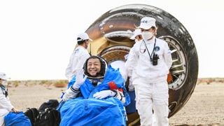 Chinese astronaut waves and smiles in reclined position with landed space capsule in background