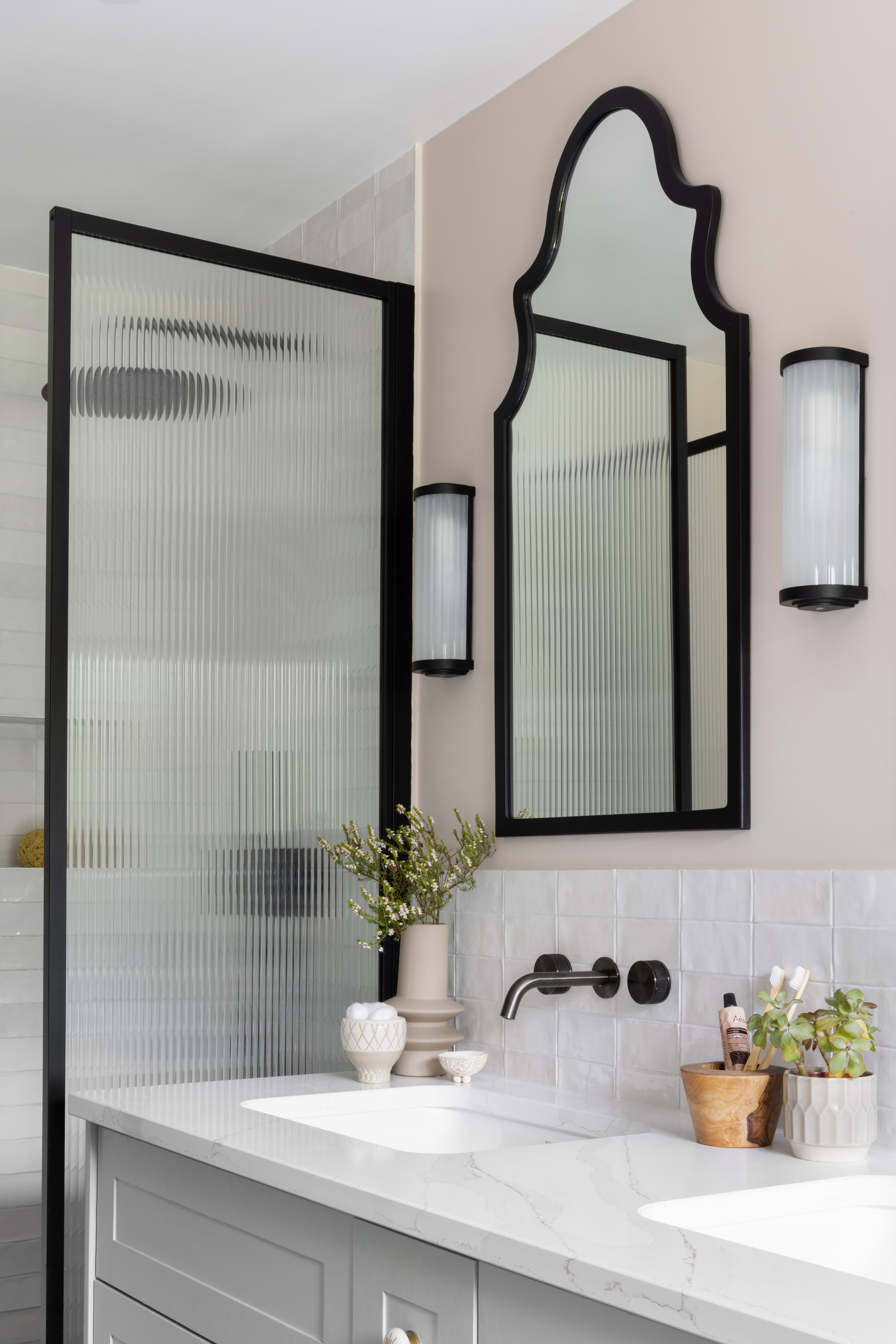 A bathroom with a double vanity, a black framed mirror, sconces, and a black framed shower panel with fluted glass